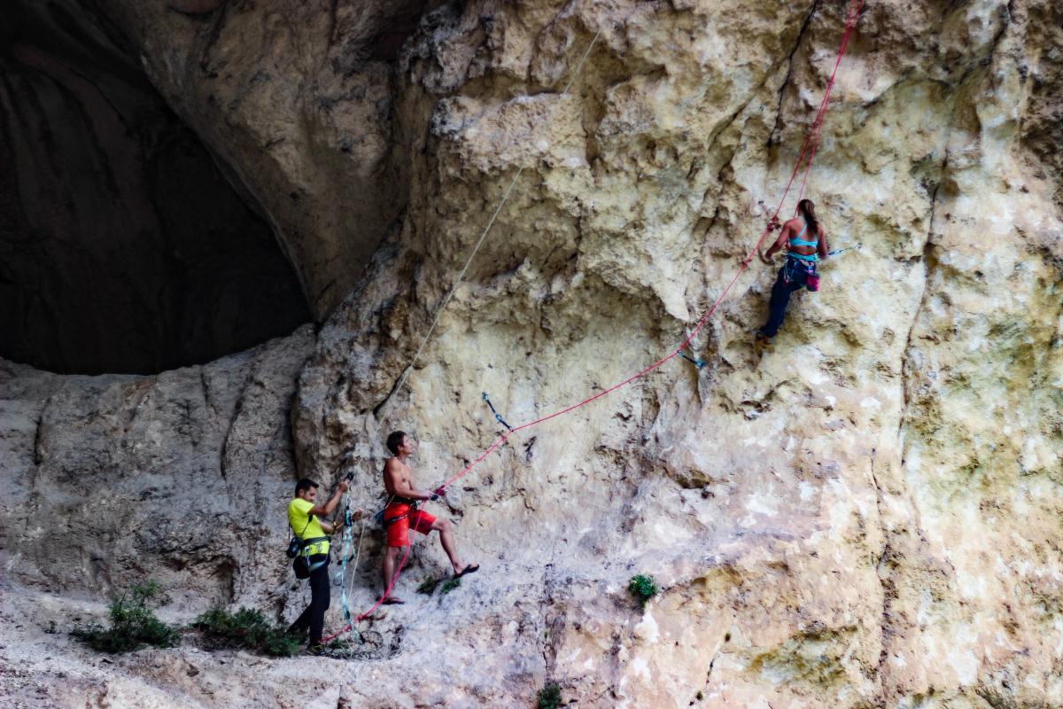 One person climbing a rock face with two people holding ropes to the lower left of the rock face